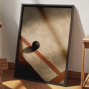 Peut inclure: Une impression d'art murale rectangulaire encadrée présentant une surface beige texturée avec une sphère sombre projetant une ombre. Deux lignes diagonales brun-rouge se croisent, créant un contraste géométrique sur le fond neutre.