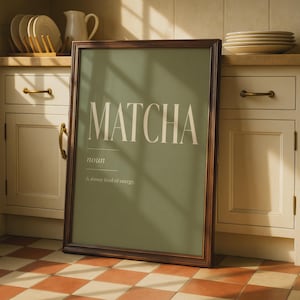 May include: A framed wall art print featuring a sage green background with the white text MATCHA, the word noun, and the phrase A slower kind of energy. The print is enclosed in a dark brown wooden frame and stands upright on a tiled floor.