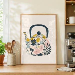 Puede incluir: Un cuadro enmarcado con un diseño de tetera floral. La tetera está adornada con flores coloridas en tonos amarillos, rosas, azules y azul marino sobre un fondo crema. El marco es de madera clara, añadiendo un toque cálido a la decoración de la cocina.
