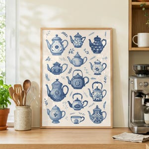 Può includere: Una stampa incorniciata con una collezione di teiere blu e bianche in vari design. Le teiere sono disposte su uno sfondo color crema con accenti botanici. La stampa è esposta su un piano di lavoro in legno vicino a una finestra.