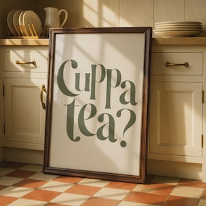 Puede incluir: Un cuadro rectangular con marco de madera marrón oscuro que presenta el texto "Cuppa tea?" en letras grandes de estilo serif en color verde salvia sobre un fondo crema, exhibido en una cocina con armarios y vajilla.
