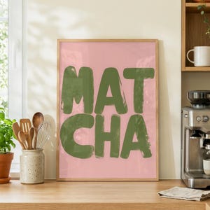 May include: A framed print featuring the word "MATCHA" in large, olive green brushstrokes against a pink backdrop. The wooden frame complements the design. The print is displayed on a wooden countertop, with kitchen utensils and a coffee machine visible.
