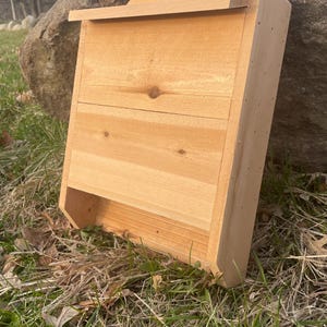 May include: A wooden bat house with a rectangular design. The bat house is made of light-colored wood and features a small entrance at the bottom. It is designed to provide shelter for bats and is suitable for outdoor use.