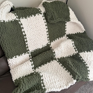 Può includere: Una coperta fatta a mano, a maglia grossa, con un motivo a scacchiera. La coperta presenta quadrati alternati di filato verde oliva e color crema. La coperta è drappeggiata su un divano in pelle marrone.
