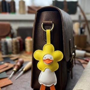 Peut inclure: Un sac en cuir marron avec un porte-clés en forme de fleur jaune. Le porte-clés représente un corps blanc, des jambes orange et une tête de fleur jaune avec un visage de canard. Le sac a un anneau en laiton et une sangle jaune.