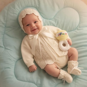 May include: A baby in a cream-coloured knitted outfit, comprising a bonnet, sweater, romper, and booties. The outfit has a cable knit pattern and satin ribbon detailing. The baby is holding a yellow and white rattle.