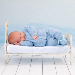May include: A sleeping baby wearing a light blue knitted outfit, including a sweater and pants, lies on a small white bed. The bed has a distressed, vintage-style metal frame. The background is a soft blue, creating a serene and calming scene.