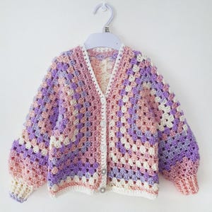 May include: A pastel-coloured, handmade crochet cardigan in shades of pink, purple, and cream, displayed on a white hanger. The cardigan features a classic granny square design, long sleeves, and button closures. The garment is likely for a baby or toddler.