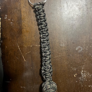 May include: A black and green paracord keychain with a silver metal ring. The keychain is made of tightly woven cord and features a decorative knot at the bottom. The keychain is designed for attaching keys or other small items.