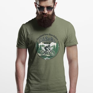 May include: Olive green t-shirt with a circular graphic showing a mountain biker, trees, and the text "Cleveland Area Slow Rollers CASERs MTB". The design uses green, white, and brown.