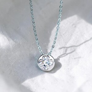 Puede incluir: Un collar de tono plateado con un colgante circular que presenta una piedra preciosa transparente en forma de flor de cuatro pétalos en el centro. El colgante cuelga de una delicada cadena de metal plateado con eslabones ovalados uniformes.
