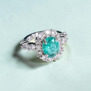 Op de afbeelding: Zilveren ring met een grote, ovaal geslepen, tealkleurige edelsteen, omringd door kleinere, heldere edelstenen. De ringband is ook versierd met heldere edelstenen, wat een halo-effect creëert. De achtergrond is lichtblauw.