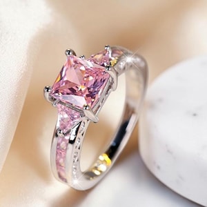 Op de afbeelding: Een zilverkleurige ring met een grote, vierkant geslepen roze steen die door vier pootjes wordt vastgehouden. De band is versierd met kleinere driehoekige roze stenen aan de zijkanten en een rij kleine roze steentjes in de ringband.