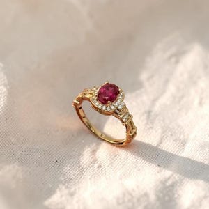 Puede incluir: Un anillo de oro con una piedra preciosa de color rosa intenso de corte ovalado en el centro, rodeada por un halo de pequeñas piedras claras. La banda está hecha de oro pulido e incluye pequeñas piedras claras adicionales en los hombros.
