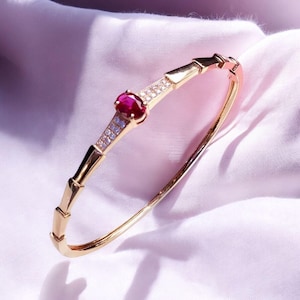 May include: A gold-colored bangle bracelet with a central ruby gemstone and small, clear accent stones. The bracelet has a segmented design with a curved shape, resting on a white fabric surface. The ruby is a deep red color.