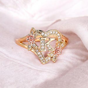 Könnte beinhalten: Ein goldfarbener Ring mit einem herzförmigen Rahmen, der mit kleinen klaren Steinen besetzt ist. In der Mitte des Herzens befindet sich der Buchstabe K, der ebenfalls mit klaren Steinen verziert ist. Zwei kleine roségoldene Blumen sind am Herz befestigt.