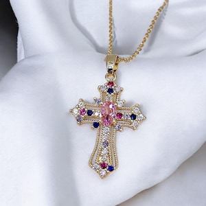 Op de afbeelding: Een gouden ketting met een kruisvormige hanger. Het kruis is versierd met een roze edelsteen in het midden en kleinere gekleurde edelstenen in roze, blauw en wit. De ketting hangt aan een fijne gouden ketting.
