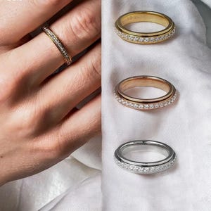 Könnte beinhalten: Drei Spinner-Ringe in Gold-, Roségold- und Silbertönen, jeweils mit einem Band, das mit kleinen klaren Steinen besetzt ist. Ein Ring wird an einem Finger getragen, während die anderen beiden separat präsentiert werden und das metallische Finish zeigen.