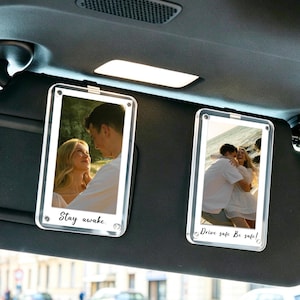 Op de afbeelding: Twee rechthoekige, doorzichtige acryl fotolijsten bevestigd aan een donkergrijze zonneklep van een auto. Elke lijst bevat een foto van een koppel. De linkerlijst bevat de tekst Stay awake. De rechterlijst bevat de tekst Drive safe. Be safe!