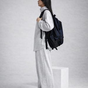 May include: A person wearing a gray sweatshirt, matching sweatpants, and white sneakers, carrying a navy blue backpack. The outfit is casual and comfortable. The background is a plain, light gray.