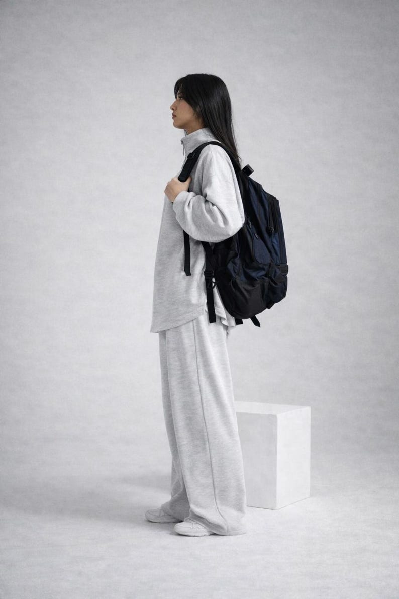 May include: A person wearing a gray sweatshirt, matching sweatpants, and white sneakers, carrying a navy blue backpack. The outfit is casual and comfortable. The background is a plain, light gray.