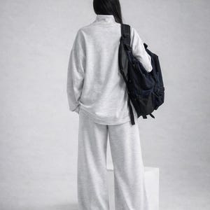 May include: A person wearing a light gray, oversized sweatshirt and matching wide-leg pants. A black backpack is worn over the shoulder. The outfit is a casual, comfortable style, suitable for everyday wear.