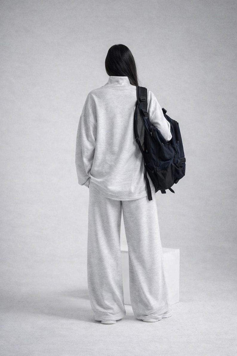 May include: A person wearing a light gray, oversized sweatshirt and matching wide-leg pants. A black backpack is worn over the shoulder. The outfit is a casual, comfortable style, suitable for everyday wear.