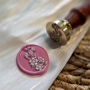 Può includere: Un sigillo di cera rosa con un disegno floreale argentato accanto a un timbro di cera con manico in oro e legno. Il sigillo è su un tessuto bianco, con un cesto di vimini sullo sfondo. Il timbro ha un motivo floreale.