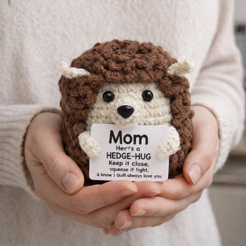 May include: A crocheted hedgehog toy with a brown body and cream-coloured face and ears. It holds a small white card with the word "Mom" and a message. The hedgehog is held in two hands. The card also includes a short message.