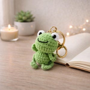 May include: A green crocheted frog keyring with a gold-coloured ring. The frog has white eyes with black pupils and a smiling mouth. The background includes a lit candle, a small potted plant, and an open book.