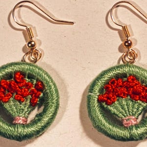 May include: Embroidered earrings with a green circular design and a red floral arrangement. The earrings have gold-coloured hooks. The floral arrangement is in a basket shape with a pink ribbon. Handmade earrings.