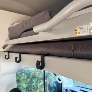 Könnte beinhalten: Innenansicht eines Wohnmobils mit einem Etagenbett. Das Bett hat eine graue Matratze und einen weißen Rahmen mit schwarzen Haken. Eine silberne Stange verläuft über die Oberseite. Ein Diagramm und ein silberner Knopf sind an der Seite sichtbar.