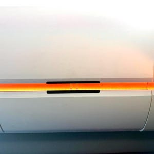 Könnte beinhalten: Ein weißes Wandklimagerät mit horizontalem Paneeldesign. Ein leuchtend orangefarbener LED-Lichtstreifen strahlt aus der mittleren Fuge, die durch einen kleinen schwarzen rechteckigen Ausschnitt in der Mitte des Geräts unterbrochen wird.