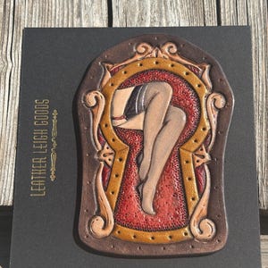 May include: Leather Leigh Goods artwork featuring a keyhole design with a woman's legs in stockings. The keyhole is framed in brown leather with gold accents, set against a red background. The artwork is on a black background.