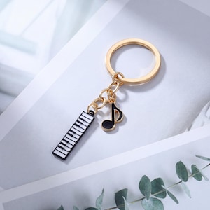May include: A gold-coloured keyring featuring a piano keyboard and a musical note charm. The piano keyboard charm is black and white, and the musical note is black. The keyring is on a white and grey background with green leaves.