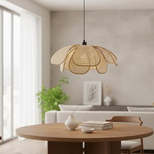 Puede incluir: Una lámpara colgante de ratán beige con forma de flor, suspendida sobre una mesa de comedor de madera. La lámpara tiene múltiples pantallas en forma de pétalo y un centro tejido. La habitación presenta una paleta de colores neutros con un sofá blanco y una planta verde.