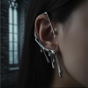 May include: A silver ear cuff with a unique, flowing design. The cuff wraps around the ear and features a series of elongated, dripping shapes, creating a fluid, artistic look. The jewelry is shown on a person's ear, with dark hair in the background.