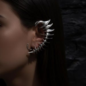 May include: A silver ear cuff featuring a unique, sculptural design. The cuff curves around the ear with a series of pointed, blade-like elements and a curved top. The jewelry is worn on the ear, creating a bold, avant-garde look.