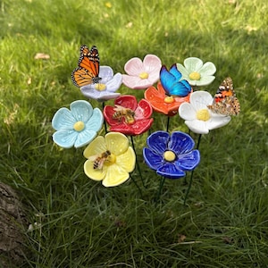 Può includere: Una collezione di picchetti da giardino a forma di fiore in ceramica in vari colori, tra cui blu, giallo, rosso, arancione, bianco e rosa. Ogni fiore presenta uno stame centrale giallo ed è decorato con una realistica figura di farfalla o ape.