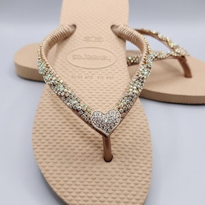 May include: Beige flip-flops featuring a textured footbed and Havaianas branding. The straps are embellished with rows of iridescent crystals and a central heart-shaped charm covered in clear rhinestones. The size is marked as USA 3/4 and EUR 35/36.
