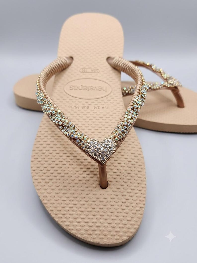May include: Beige flip-flops featuring a textured footbed and Havaianas branding. The straps are embellished with rows of iridescent crystals and a central heart-shaped charm covered in clear rhinestones. The size is marked as USA 3/4 and EUR 35/36.