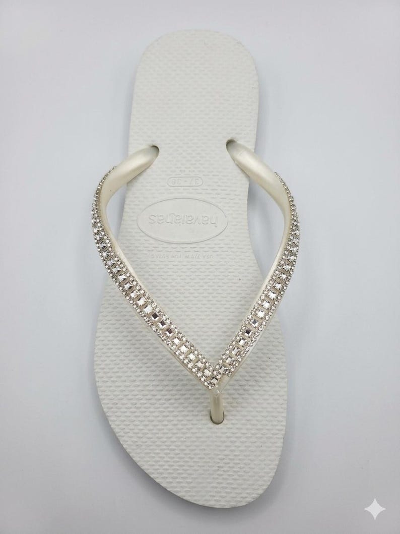 May include: White Havaianas flip-flop with a textured sole and a strap embellished with sparkling rhinestones. The flip-flop is a classic design, perfect for casual wear or beach outings.