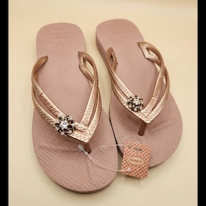 Puede incluir: Un par de chanclas rosa empolvado con correas doradas. Cada chancla presenta un adorno decorativo con pedrer&iacute;a transparente y marr&oacute;n. Una etiqueta de precio est&aacute; adherida a una de las chanclas.