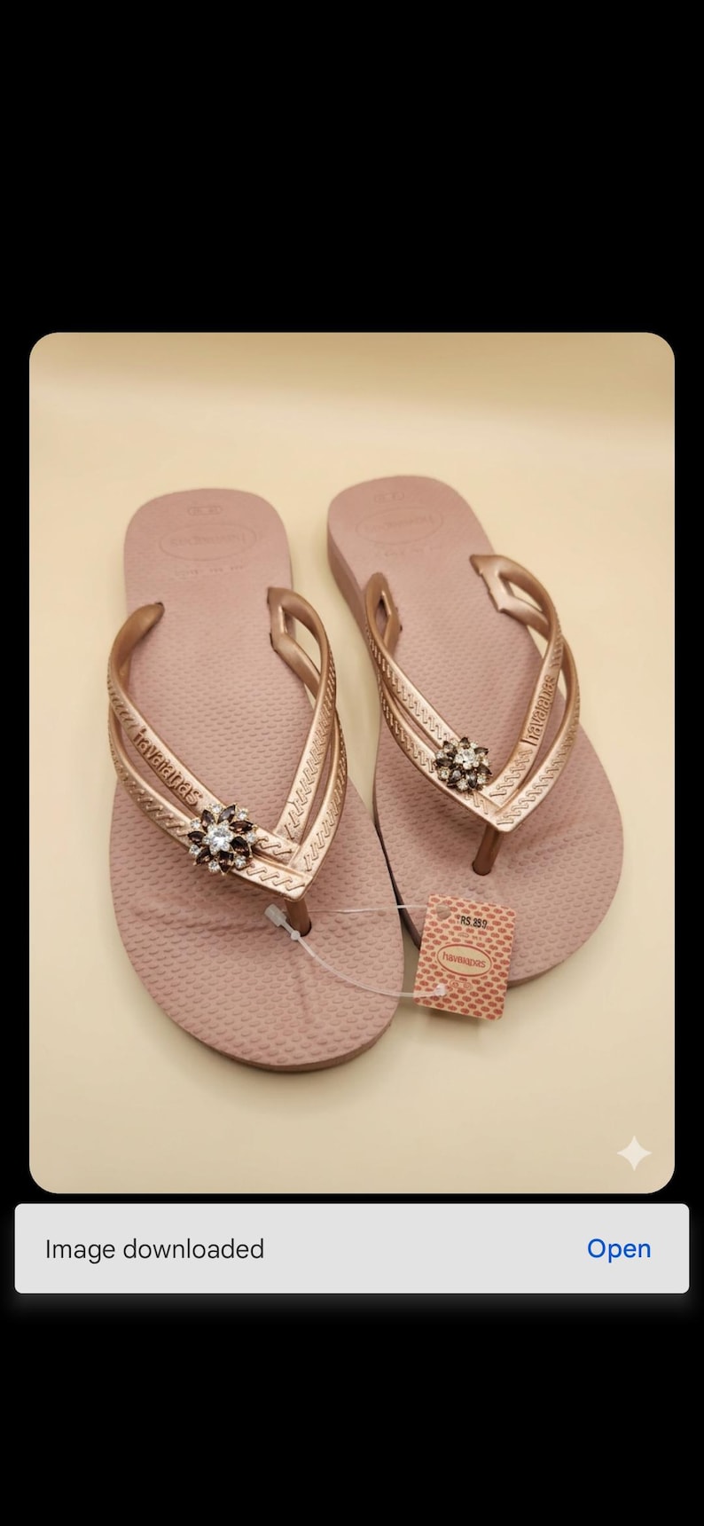 Puede incluir: Un par de chanclas rosa empolvado con correas doradas. Cada chancla presenta un adorno decorativo con pedrer&iacute;a transparente y marr&oacute;n. Una etiqueta de precio est&aacute; adherida a una de las chanclas.