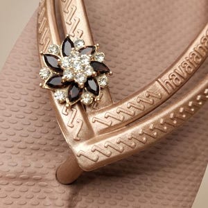 Puede incluir: Primer plano de una chancla rosa dorada con un adorno floral decorativo. La flor tiene detalles en marr&oacute;n oscuro y cristal transparente. La chancla tiene la palabra "havaianas" en la correa.