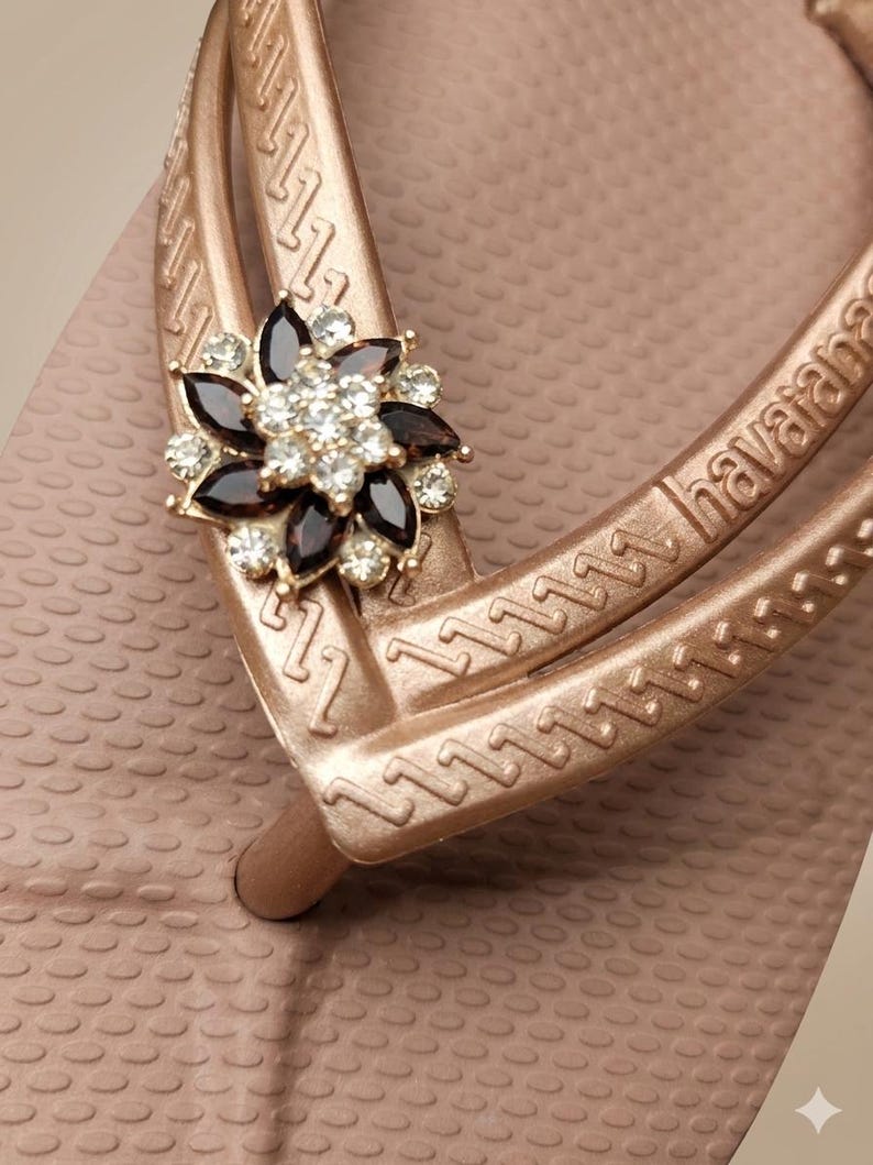 Puede incluir: Primer plano de una chancla rosa dorada con un adorno floral decorativo. La flor tiene detalles en marr&oacute;n oscuro y cristal transparente. La chancla tiene la palabra "havaianas" en la correa.