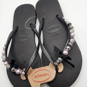 May include: A pair of black flip-flops featuring textured soles and branded straps. The straps are embellished with a row of alternating light pink pearls, clear faceted beads, and dark patterned beads, finished with a small black ribbon bow.