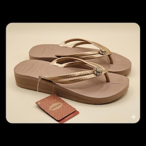 Puede incluir: Un par de chanclas Havaianas marrones con plataforma, con correas doradas y una joya decorativa. Las chanclas tienen una suela texturizada y una etiqueta adjunta. El fondo es de color beige claro.