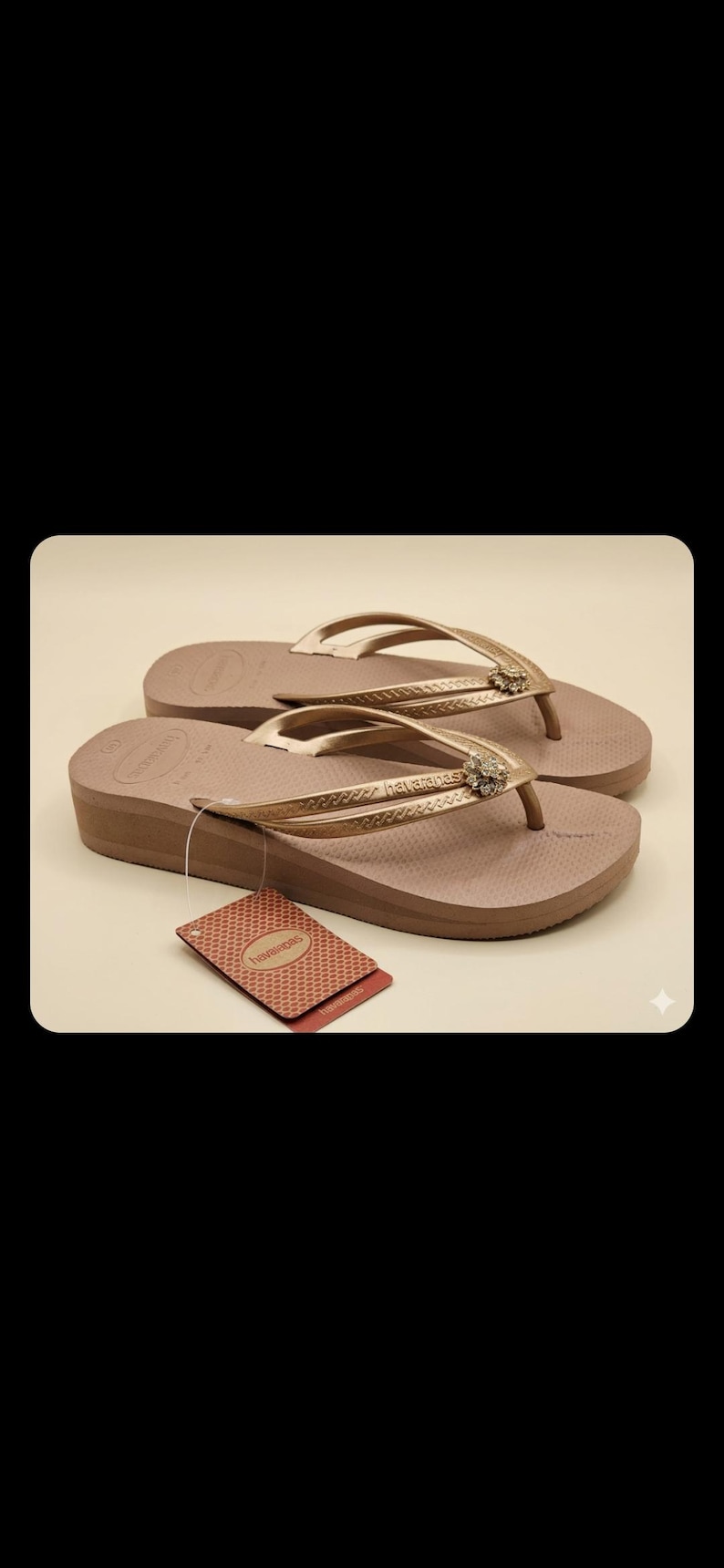 Puede incluir: Un par de chanclas Havaianas marrones con plataforma, con correas doradas y una joya decorativa. Las chanclas tienen una suela texturizada y una etiqueta adjunta. El fondo es de color beige claro.