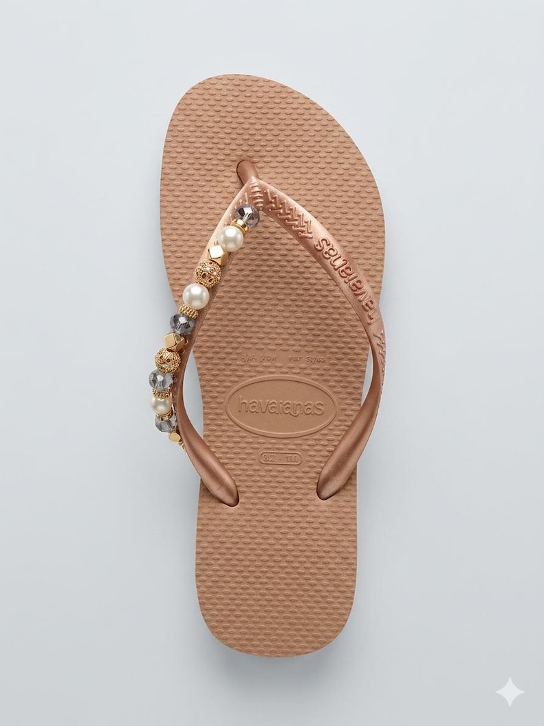 Puede incluir: Una chancla Havaianas de color oro rosa con una correa de cuentas. La correa est&aacute; adornada con cuentas doradas, perladas y cristalinas. La chancla tiene una plantilla texturizada y el logotipo de Havaianas es visible.